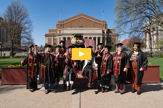 Group of graduates and Goldy on the Mall. A gold video play button in the center.